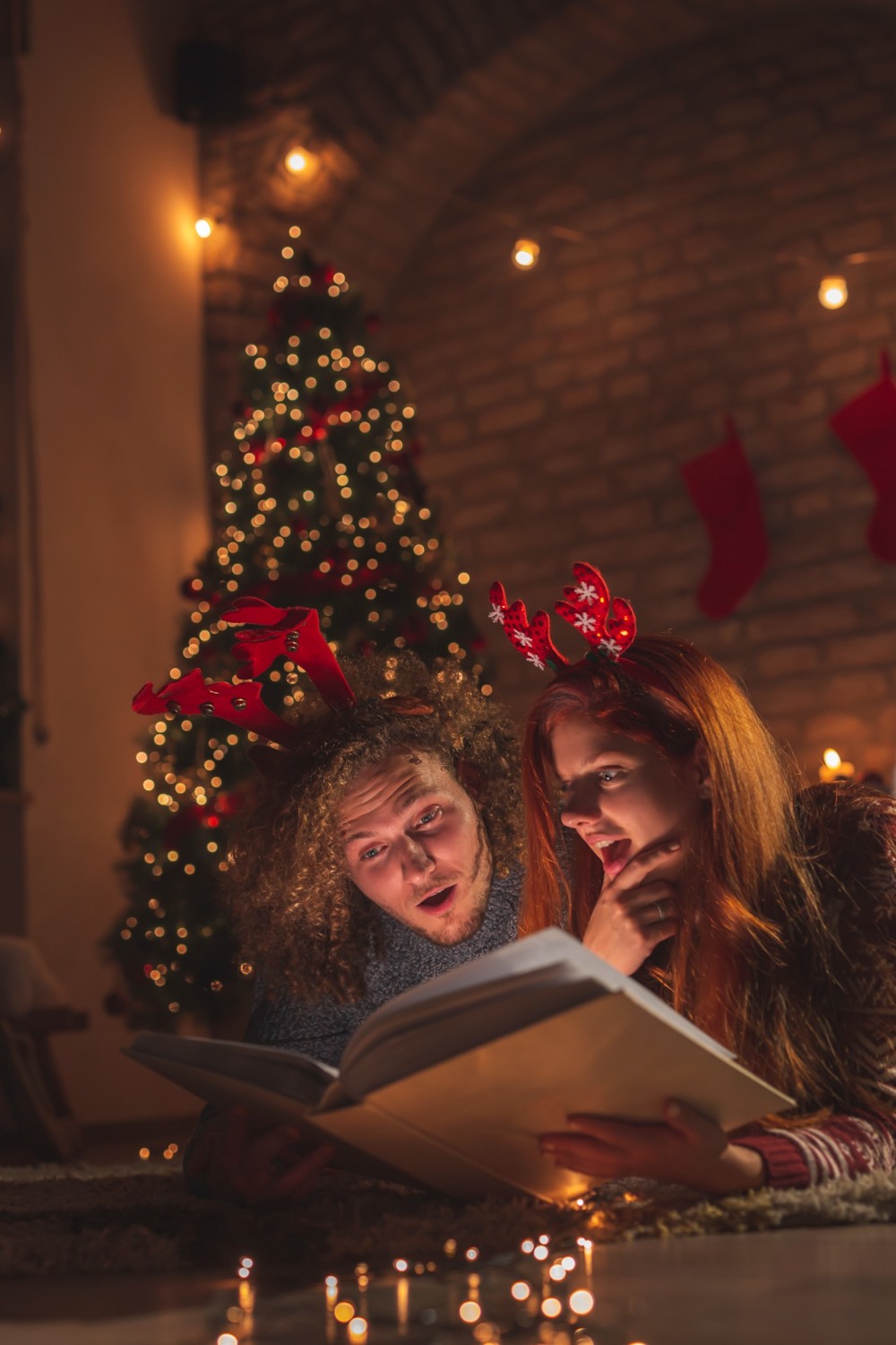 A couple reading a book together beside a Christmas tree and warm fairy lights