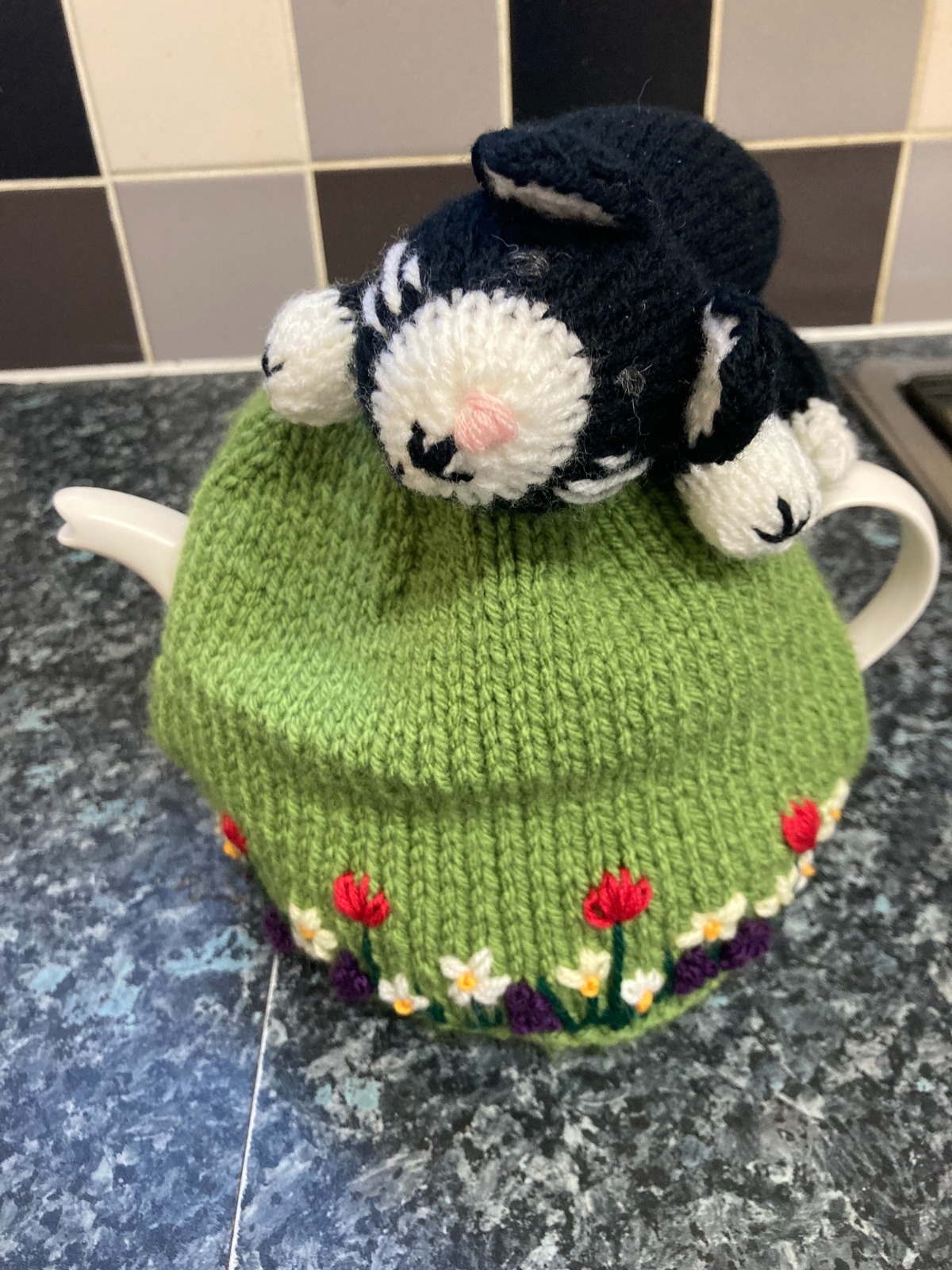 Green knitted tea cosy with embroidered flowers and a black and white cat on top